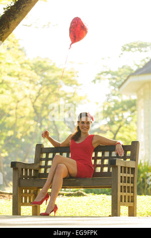 Frau mit roter Herzballon Form auf Parkbank sitzen Stockfoto