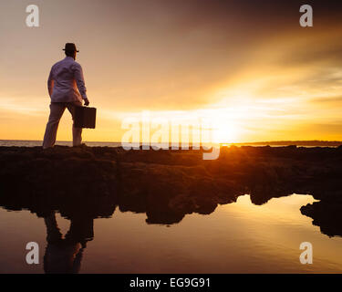 Mann, der bei Sonnenuntergang am Strand spazieren ging und einen Koffer trug Stockfoto