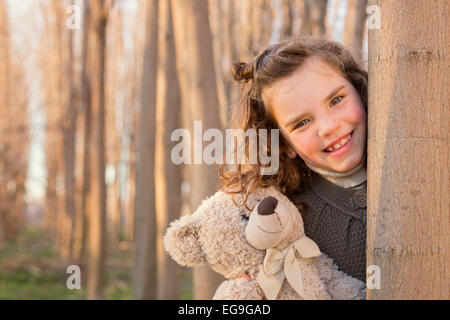 Mädchen versteckt sich hinter Baumstamm mit ihr Teddy Bär Stockfoto