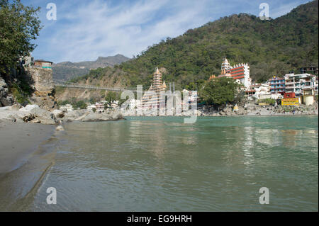 Eine Ansicht vom Strand mit den Tempeln und Bergen von Rishikesh mit Lakshman Jhula Brücke über den Fluss Ganges oder Ganga. Stockfoto