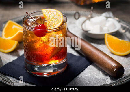 Old Fashioned Cocktail in Rocks Glass mit Eis, Orangen und einer Kirsche mit Bar-Tools im dunklen Hintergrund Stockfoto