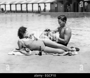 1930S 1940S PAAR TRAGEN BADEANZÜGE SITZEN ENTSPANNENDEN POOL SEITE BIER TRINKEN Stockfoto