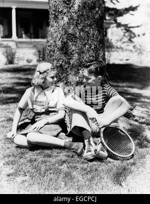 1930S 1940S JUNGE UND BLONDE MÄDCHEN SITZEN AUßERHALB VON BAUMSTAMM SPRECHEN HOLDING TENNISSCHLÄGER Stockfoto