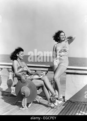 1930S 1940S DECK ZWEI FRAUEN SITZEN AUF HOTEL AM STRAND IN EINEM STÜCK BADEANZUG MODE FLORIDA USA Stockfoto