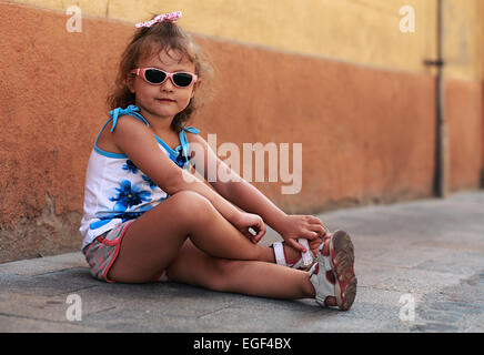 Kind Mädchenmode in Sonnenbrille auf Straße in der Nähe der Wand sitzen und Lächeln Stockfoto