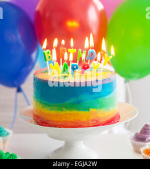 Regenbogen Kuchen und Cupcakes mit Geburtstagskerzen dekoriert. Luftballons auf dem Hintergrund Stockfoto