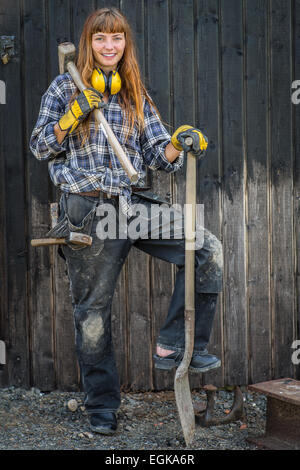 Mädchen im Bau Arbeiter Kleidung stehend mit einer Schaufel und Hammer vor der Scheune Stockfoto