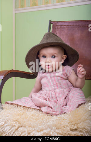 niedliche Baby tragen ein langes Kleid und Mutters Hut. Sitzend auf einem Stuhl im Zimmer Stockfoto