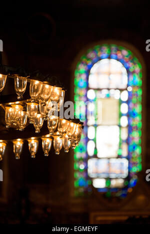 ISTANBUL, Türkei – Ein traditioneller osmanischer Kronleuchter hängt vor einem reich verzierten Buntglasfenster in der Hagia Sophia. Diese farbigen Glasfenster, die während der osmanischen Renovierung hinzugefügt wurden, schaffen markante Lichtmuster innerhalb des heiligen Raumes. Die Kombination von natürlichen und künstlichen Lichtquellen stellt ein charakteristisches Merkmal der osmanischen Moschee-Architektur dar. Stockfoto