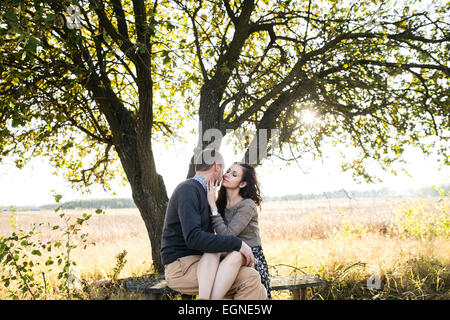 Bild eines jungen Paares küssen beim Sitzen auf einer Bank unter Baum bei Sonnenuntergang im Herbst Stockfoto