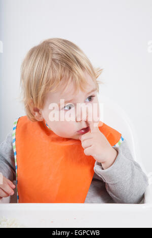 blonde kaukasische Baby siebzehn Monate Alter Orange bib grauen Pullover sitzen auf weißen Hochstuhl mit Finger-picking Nase Essen Stockfoto