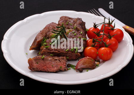 Rib-Eye Steak, Cherry-Tomaten, Knoblauch, Kräuter auf schwarzem Hintergrund Stockfoto