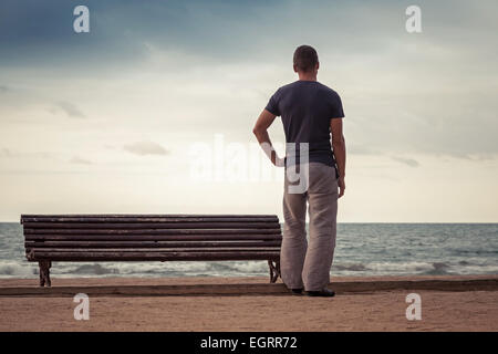 Junger Mann steht in der Nähe der alten Holzbank an der Meeresküste. Vintage getönten Foto mit Instagram Retro-toning Filterwirkung Stockfoto