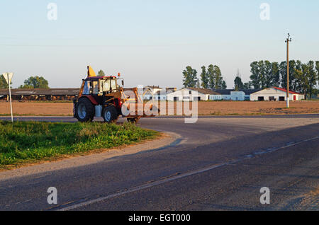 Landwirtschaftliche Zugmaschine-Bagger auf der Straße in der Nähe des Dorfes. Am Abend Sonne. Stockfoto