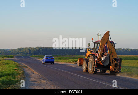 Landwirtschaftliche Zugmaschine-Bagger auf der Straße in der Nähe des Dorfes. Am Abend Sonne. Stockfoto