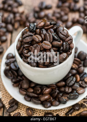 Vertikale Foto von gerösteten Kaffeebohnen in einem kleinen weißen Espressotasse und Teller auf einem rustikalen hölzernen Hintergrund hautnah. Stockfoto