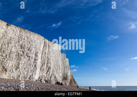 Tagesausflügler genießen Sie die Wintersonne auf dem Kiesstrand unterhalb der ersten der sieben Schwestern Kreidefelsen im Cuckmere Haven. Stockfoto