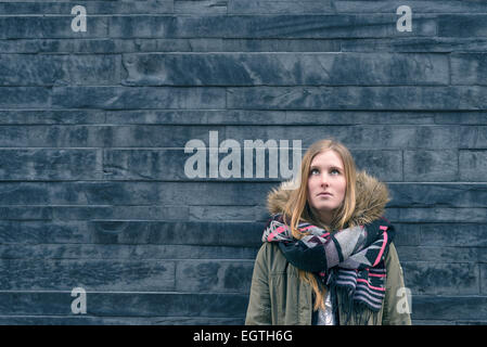 Trendige junge Studentin in warmen Winter Mode stehen denken oder Tagträumen gelehnt eine strukturierte graue Wand suchen Stockfoto