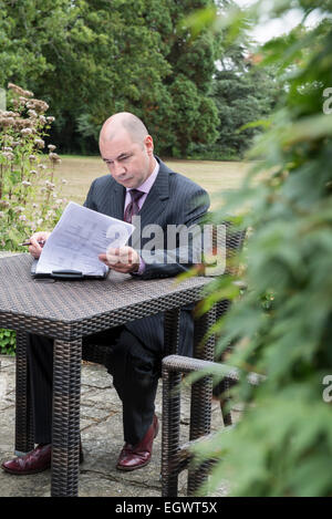 eine entspannte professionelle Unternehmer / Manager sitzt an einem Tisch draußen arbeiten mit einer Tasse Kaffee in einem Anzug an einem schönen Tag Stockfoto