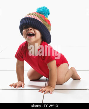 Junge (16 Monate) kniend am Boden lachen, Weste und große gestrickte Pudelmütze tragen Stockfoto