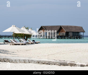Liegestühle und Sonnenschirme am Strand auf einer Inselresort auf den Malediven mit einer der Villen in der Ferne Stockfoto