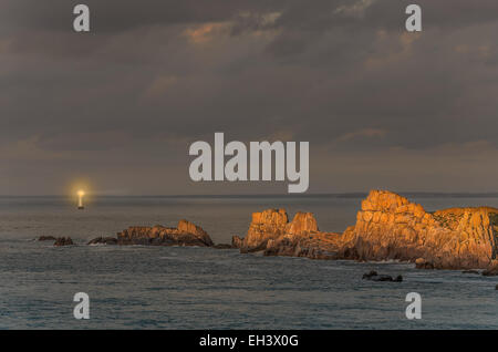 Klippen mit Leuchtturm Strahl auf Twilight, mit Felsen beleuchtet durch die letzten Sonnenstrahlen vor Sonnenuntergang, Sonnenlicht, Bretagne, Frankreich Stockfoto