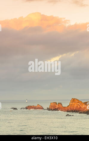 Klippen unter großen bewölkten Himmel mit Leuchtturm in Ferne Felsen beleuchtet durch die letzten Sonnenstrahlen vor Sonnenuntergang, Sonnenlicht, Saint Malo, Stockfoto