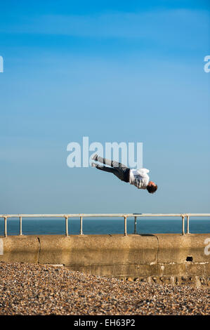 Ein junger Mann dreht in der Luft, während eine Gruppe von jungen Menschen machen das Beste aus dem sonnigen Frühlingswetter praktizieren die Kunst des Parkour, frei springen und Saltos, aus einem Steg auf Brighton Seafront mit dem Dis verwendete West Pier im Hintergrund zu tun. Brighton, East Sussex, England, UK. Stockfoto