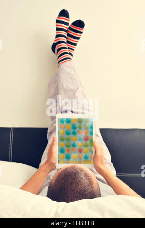 ein junger Mann im Schlafanzug im Bett mit einem Tablet mit den Füßen gegen die Wand Stockfoto