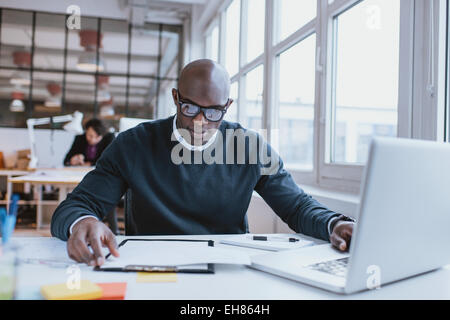 Junge afrikanische Führungskraft sitzt an seinem Schreibtisch mit Laptop Lesen eines Dokuments. Afrikaner, die im Büro arbeiten. Stockfoto