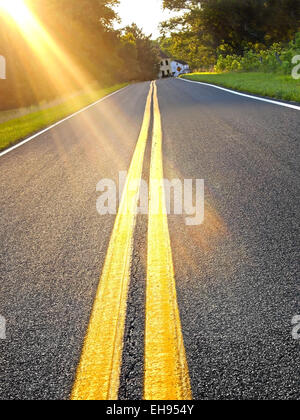Brilliant rays of late day sunlight glow across a long road through the countryside on a summer afternoon. Stockfoto