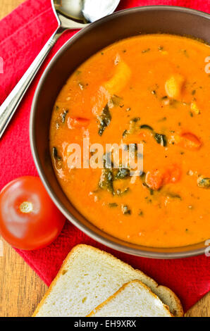 Eine Schüssel mit Gemüsesuppe mit Brot, Tomaten und Löffel auf einem Holztisch. Stockfoto