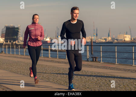 Aktive junge Paar läuft auf eine Strandpromenade mit der sportlichen Frau den Weg nähert sich die Kamera in eine Heilung zu passen Stockfoto