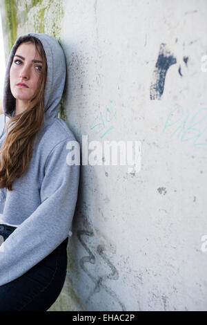 Eine junge 13 14 15 jährige Teenager-Mädchen im freien draußen allein durch sich selbst trägt eine Greey hoody Hoodie Jacke vor einer schmutzigen Betonwand UK Stockfoto