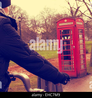 ein junger Mann, mit dem Fahrrad im Hyde Park im Winter in London, Vereinigtes Königreich, mit einer typischen roten Telefonzelle in der Hinterg Stockfoto