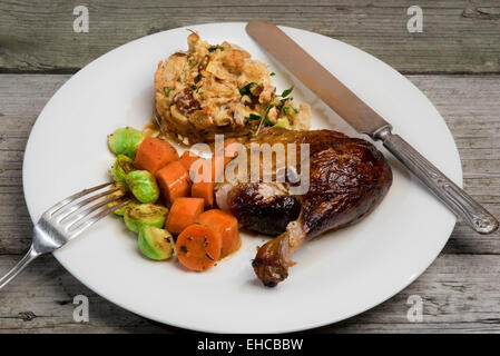 Gebratene Entenkeule mit Apfel und Kräuter Füllung, Möhren und Rosenkohl. Serviert auf einem einfachen weißen Teller auf rustikal verwittert Stockfoto