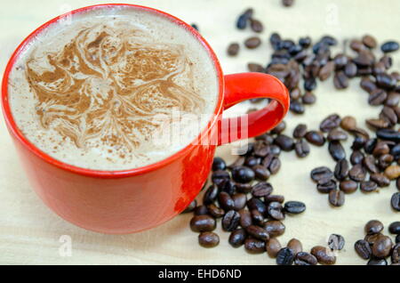 Rote Tasse Kaffee mit dekorierten Schaum und Kaffeebohnen auf Holztisch Stockfoto