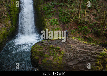 männlichen Fotografen stehen auf einem großen Felsen direkt neben einem massiven Wasserfall in Brautmoden fällt Oregon Überprüfung seiner Aufnahmen Stockfoto