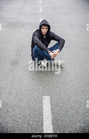 Hübscher junger Mann in schwarzer Hoodie Pullover sitzt in der Mitte der Straße, Blick in die Kamera Stockfoto