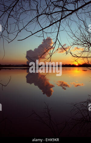 Ein Kraftwerk Freigabe Schwaden von Wasserdampf in den Himmel bei Sonnenuntergang, spiegelt sich in der Oberfläche des großen Teiches. Stockfoto