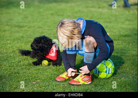 Ein Junge sitzt auf seinem Fußball und sich seine Schnürsenkel auf seine g = Fußballschuhe während seinem Hund sitzt neben ihm ein Halstuch Sport Stockfoto