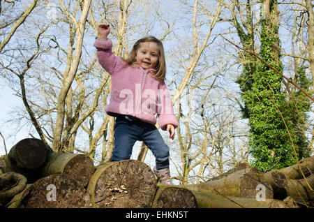 Mädchen-zwei-und-ein-halb Jahre alt, Klettern über einen Stapel der Protokolle in einem Wald im Winter, Sussex, UK. März. Stockfoto