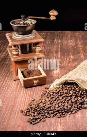 Kaffeebohnen-Ly verschüttet aus einem hessischen Beutel auf einem Holztisch mit einer alten Mühle in den Rücken. Stockfoto