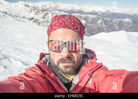 Erwachsenen europäischen Mann mit Bart, Sonnenbrille und Hut, unter Selfie auf verschneiten Hang mit schönen schneebedeckten Alpen. Stockfoto