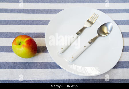 White Plate mit Edelstahl-Löffel und Gabel setzen neben Red und Green Apple auf blauem Hintergrund Stockfoto