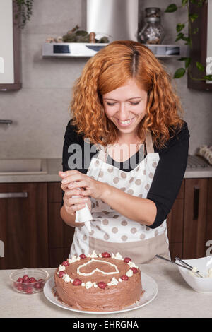 Glücklich rothaarige Frau Schokoladenkuchen mit Liebe Herzen, in der Küche zu Hause dekorieren. Stockfoto