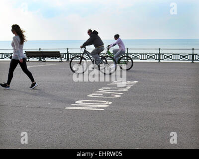 Zwei männliche Radfahrer, nicht tragen von Sturzhelmen, ignorieren Nein Radfahren Zeichen gemalt auf dem Rollfeld beim Radfahren und Blick auf ein hübsches Mädchen an Hove Promenade in der Stadt von Brighton and Hove, East Sussex Stockfoto