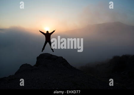 Die Frau springt und hebt ihre Arme in den Sieg. Sie betrachtet den wunderschönen Sonnenuntergang in Bergen. Stockfoto