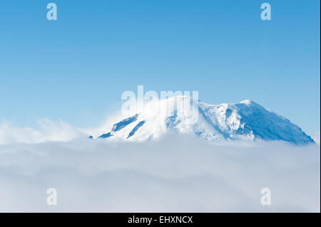 Luftaufnahme von Mt. Rainier peeking über eine Wolkendecke mit klarem, blauem Himmel und Wind peitschen Schnee vom Gipfel Stockfoto