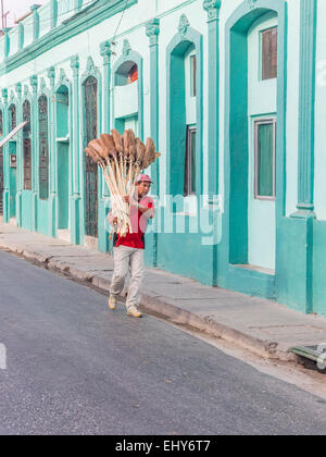 Hispanic Besen Verkäufer trägt über ein Dutzend Besen, wie er in den Straßen von Santiago De Cuba, vorbei an den bunten Fassaden geht Stockfoto